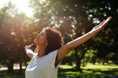 Young woman standing with her eyes closed in a park and raising her arms to the sky on a sunny summer day